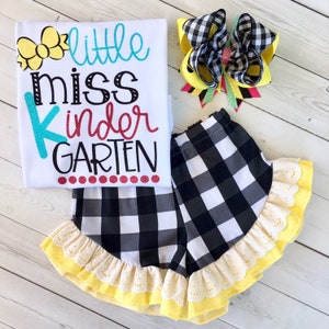 Pu&ograve; includere: Una maglietta bianca con il testo "little miss kinder GARDEN" in lettere colorate. La maglietta &egrave; abbinata a pantaloncini a quadri neri e bianchi con un bordo in pizzo giallo e bianco. &Egrave; incluso anche un fiocco a quadri giallo e nero.