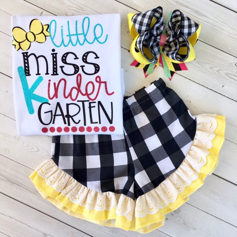 Pu&ograve; includere: Una maglietta bianca con il testo "little miss kinder GARDEN" in lettere colorate. La maglietta &egrave; abbinata a pantaloncini a quadri neri e bianchi con un bordo in pizzo giallo e bianco. &Egrave; incluso anche un fiocco a quadri giallo e nero.