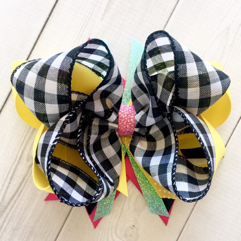 Pu&ograve; includere: Un grande fiocco per capelli a scacchi neri e bianchi con accenti di nastro giallo, verde e rosa. Il fiocco &egrave; legato con un nastro rosa scintillante al centro.