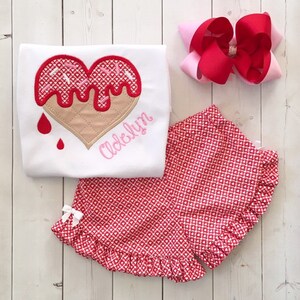 May include: A white t-shirt with a red and pink heart-shaped ice cream cone design and the name "Adeklyn" embroidered on the front. The shirt is paired with red and white gingham shorts with a ruffled hem.
