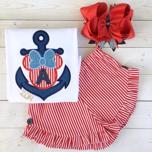 May include: A white t-shirt with a blue and red anchor embroidered on it. The anchor has a red and white striped bow and the letter "A" in the center. The shirt also has the year "2019" embroidered on it. A red and white striped skirt with a ruffle trim. A red bow with a silver glitter accent.