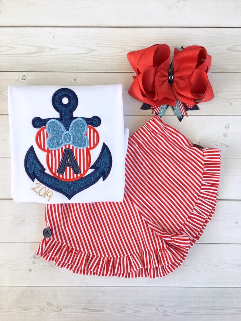 May include: A white t-shirt with a blue and red anchor embroidered on it. The anchor has a red and white striped bow and the letter "A" in the center. The shirt also has the year "2019" embroidered on it. A red and white striped skirt with a ruffle trim. A red bow with a silver glitter accent.