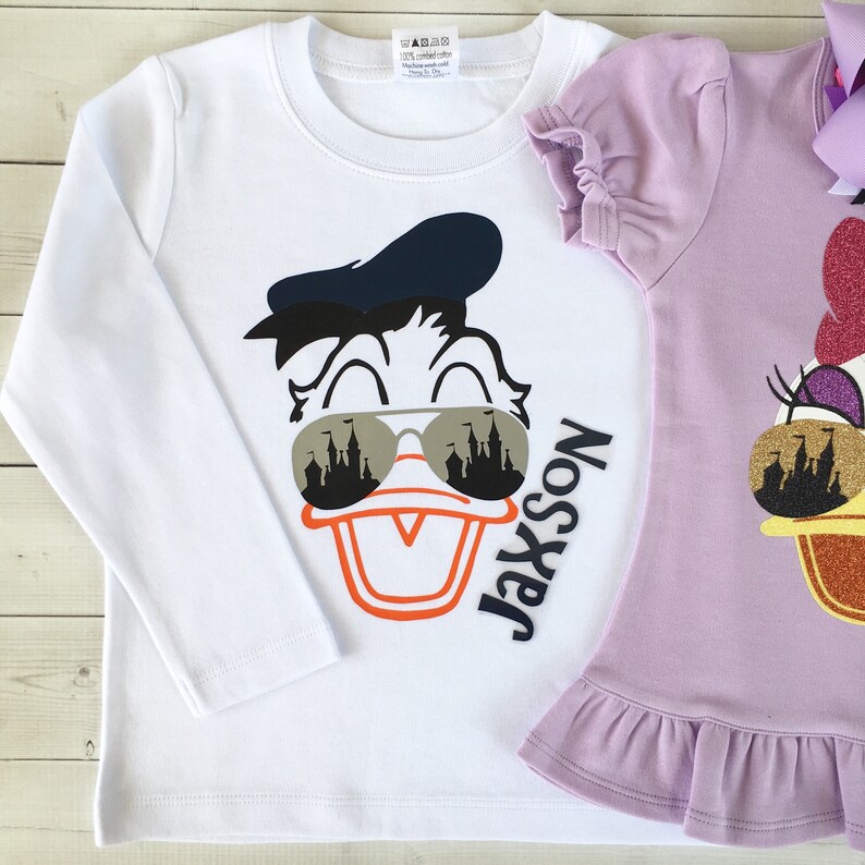 Puede incluir: Camiseta blanca de manga larga con una imagen de dibujos animados de Donald Duck con gafas de sol con un reflejo de castillo. El texto "Jaxson" est&aacute; escrito verticalmente en la camiseta.