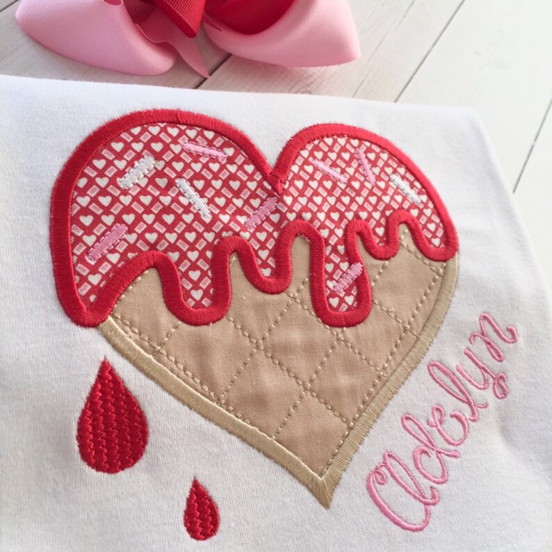 May include: A white t-shirt with a red and pink heart-shaped ice cream cone design. The ice cream is dripping and has pink sprinkles. The name "Acloelyn" is embroidered in pink below the ice cream.