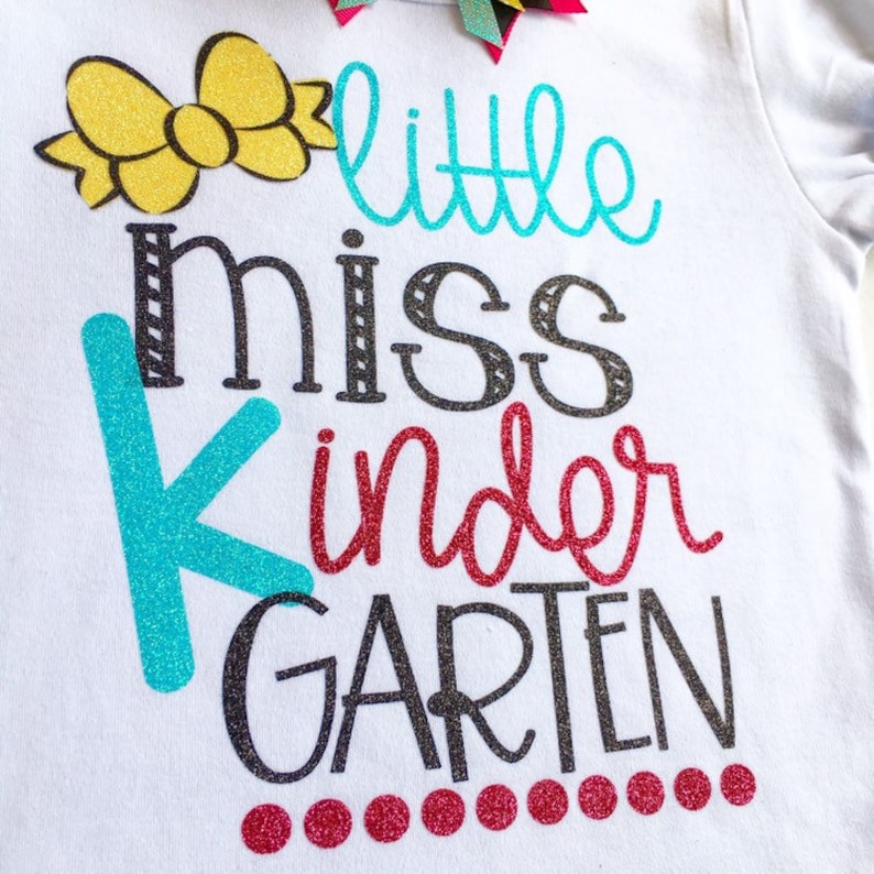 Pu&ograve; includere: Maglietta bianca con un fiocco giallo, scritta in glitter turchese, rosso e nero che dice "little miss kindergarten".