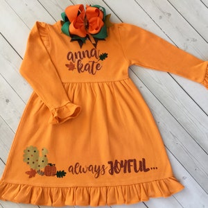 May include: Orange long-sleeved dress with a ruffled hem and cuffs. The dress features the text "anna kate" and "always JOYFUL..." in a glittery font, along with a squirrel and pumpkin graphic. A large orange and green bow adorns the neckline.