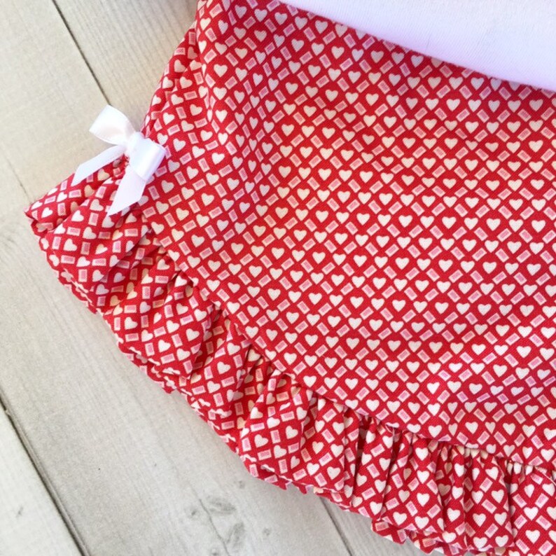 May include: Red fabric with a white heart pattern and a white ruffle trim. The fabric has a white bow on the left side.