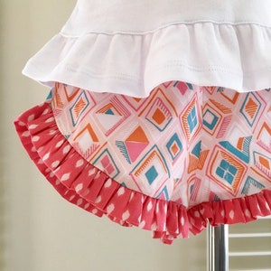 K&ouml;nnte beinhalten: Wei&szlig;es Oberteil mit R&uuml;schen und einem rosa-blauen, geometrisch gemusterten R&uuml;schenrock. Der Rock hat einen wei&szlig;en Hintergrund mit rosa, orangefarbenen und blauen geometrischen Formen.