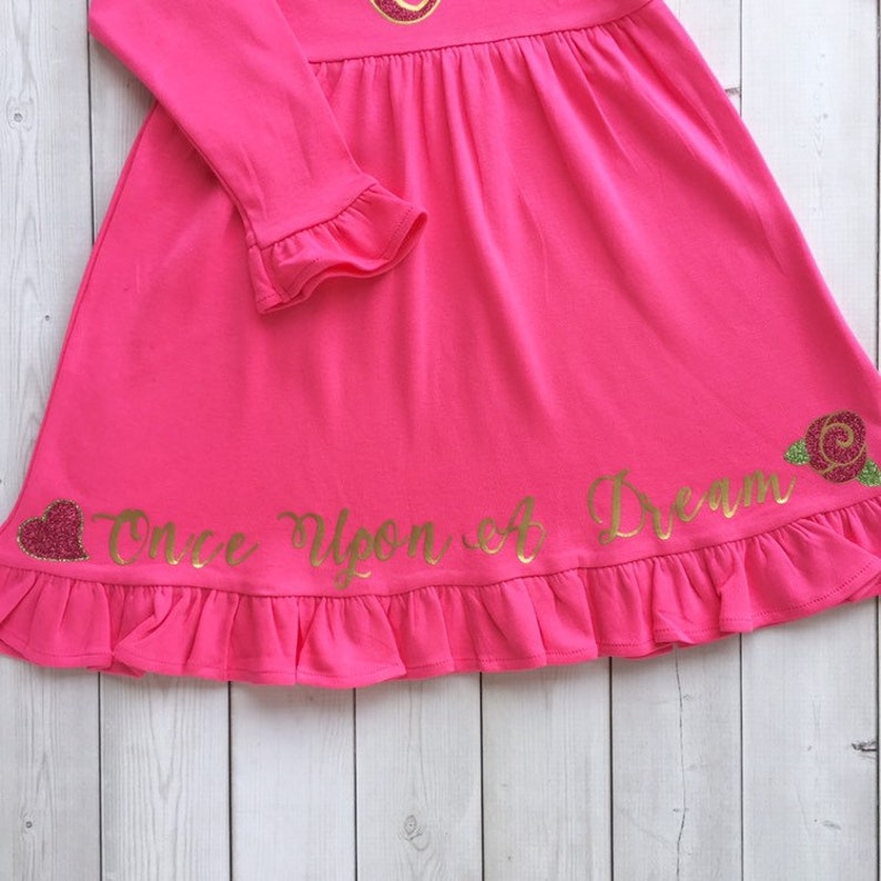 Peut inclure: Une robe rose &agrave; manches longues avec un ourlet &agrave; volants. La robe est orn&eacute;e d'un lettrage paillet&eacute; dor&eacute; qui dit "Once Upon a Dream" et d'un c&oelig;ur et d'une rose paillet&eacute;s dor&eacute;s en bas.