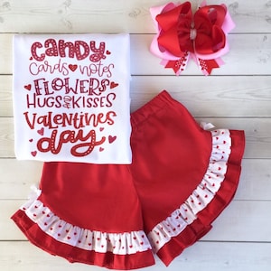 May include: Red and white Valentine's Day outfit for a girl. The outfit includes a white t-shirt with the text "candy. cards notes flowers hugs&kisses valentines day" in red glitter, and red shorts with white ruffle trim and white hearts.