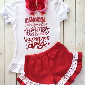 May include: A white t-shirt with a red glitter design that says "candy cards notes flowers hugs kisses valentines day". The shirt is paired with red shorts with white polka dots and a white ruffle trim.