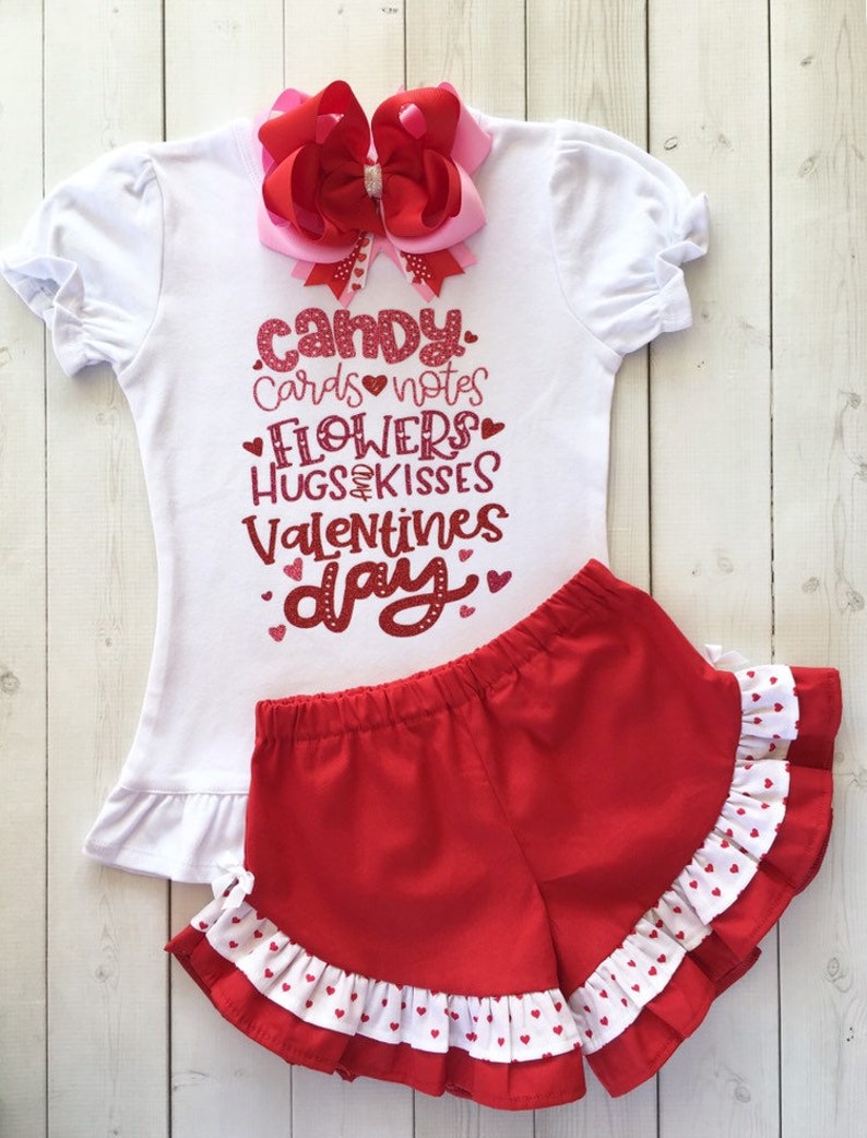 May include: A white t-shirt with a red glitter design that says "candy cards notes flowers hugs kisses valentines day". The shirt is paired with red shorts with white polka dots and a white ruffle trim.