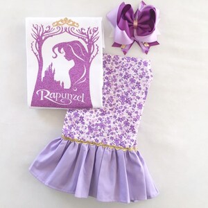 May include: A purple and white floral print dress with a ruffled hem and a gold trim. The dress is paired with a white t-shirt featuring a purple silhouette of Rapunzel with a gold crown and the word "Rapunzel" in gold glitter. A purple bow with a gold center is also included.
