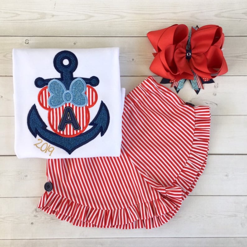 May include: A white t-shirt with a blue and red anchor design featuring a Minnie Mouse head with a bow and the letter A. The year 2019 is embroidered below the anchor. A red and white striped skirt with a ruffle hem and a red bow are also pictured.