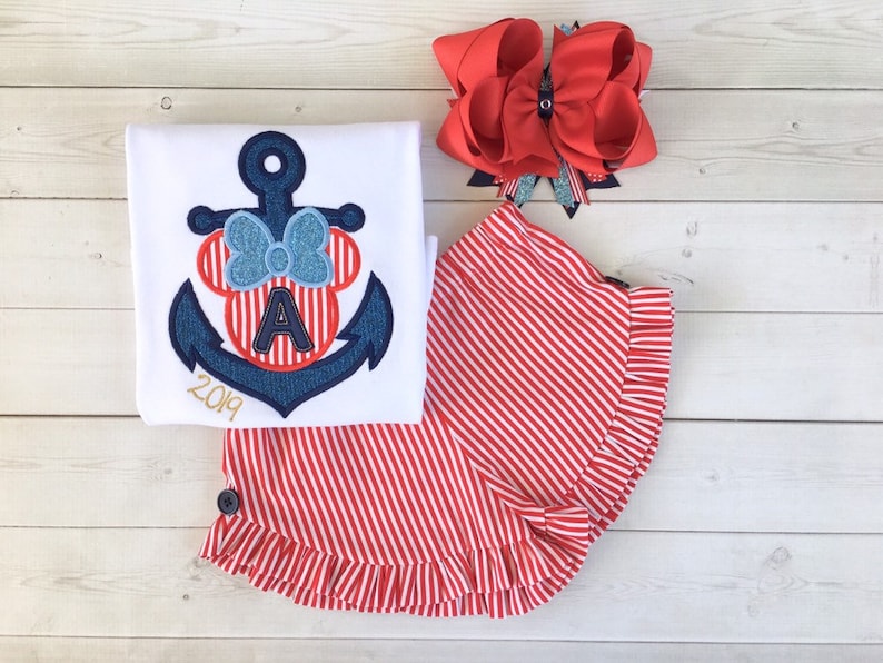 May include: A white t-shirt with a blue anchor and red Minnie Mouse head with a blue bow and the letter "A" in the middle. The shirt also has the year "2019" embroidered on it. A red and white striped skirt with a ruffle trim. A red bow with a silver glitter accent.