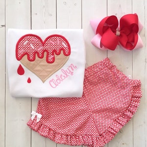 May include: A white t-shirt with a pink and red heart-shaped ice cream cone design and the name "Adelyn" embroidered on the bottom. The shirt is paired with red and white heart-patterned shorts with a white ruffle.