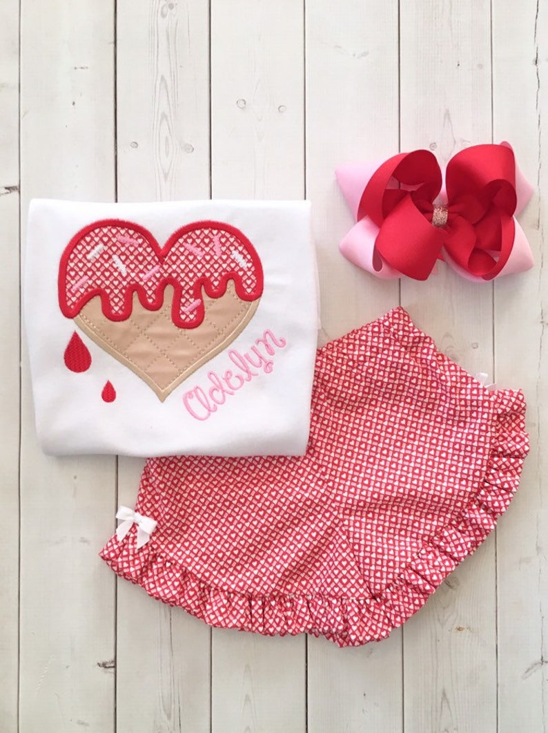 May include: A white t-shirt with a pink and red heart-shaped ice cream cone design and the name "Adelyn" embroidered on the bottom. The shirt is paired with red and white heart-patterned shorts with a white ruffle.