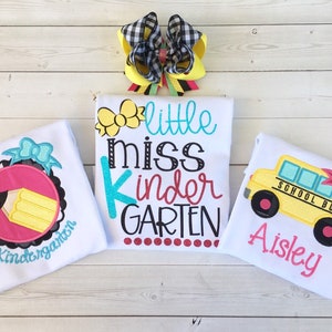 Pu&ograve; includere: Tre magliette bianche con disegni ricamati per un bambino. La prima maglietta ha un cerchio rosa con una matita gialla e le parole "Kindergarten". La seconda maglietta ha fiori gialli e le parole "little miss kinder GARDEN". La terza maglietta ha un autobus scolastico giallo con un fiocco rosa e le parole "SCHOOL BUS" e "Aisley".