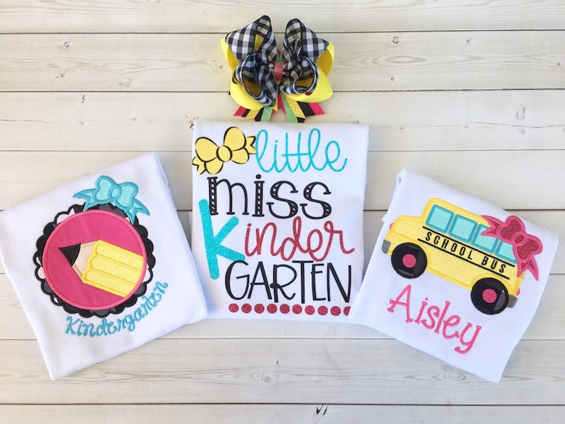 Pu&ograve; includere: Tre magliette bianche con disegni ricamati per un bambino. La prima maglietta ha un cerchio rosa con una matita gialla e le parole "Kindergarten". La seconda maglietta ha fiori gialli e le parole "little miss kinder GARDEN". La terza maglietta ha un autobus scolastico giallo con un fiocco rosa e le parole "SCHOOL BUS" e "Aisley".