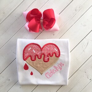 May include: White t-shirt with a red heart-shaped ice cream cone with pink sprinkles and the name "Ackerlyn" embroidered in pink.