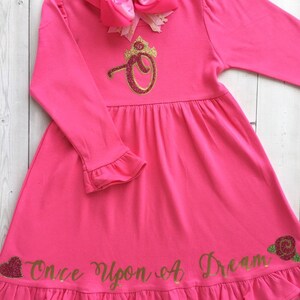 Peut inclure: Robe longue &agrave; manches longues rose avec une couronne dor&eacute;e &agrave; paillettes et le texte "Once Upon A Dream" en bas. La robe a un ourlet et des manches &agrave; volants.