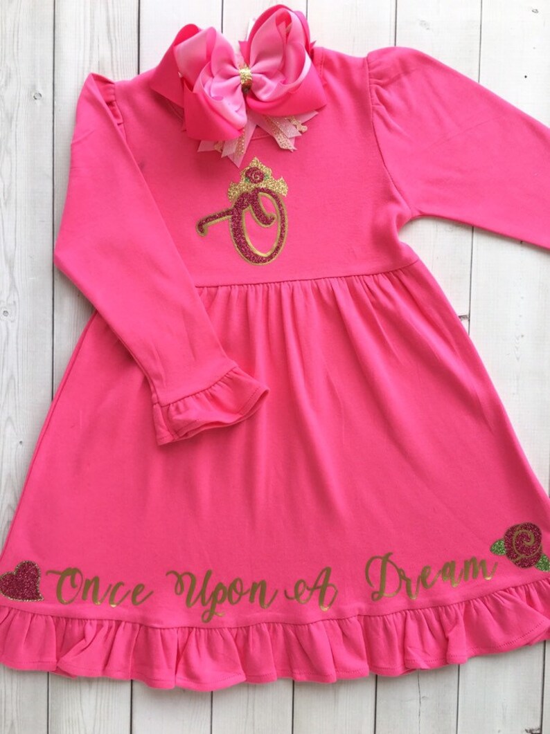 Peut inclure: Robe longue &agrave; manches longues rose avec une couronne dor&eacute;e &agrave; paillettes et le texte "Once Upon A Dream" en bas. La robe a un ourlet et des manches &agrave; volants.