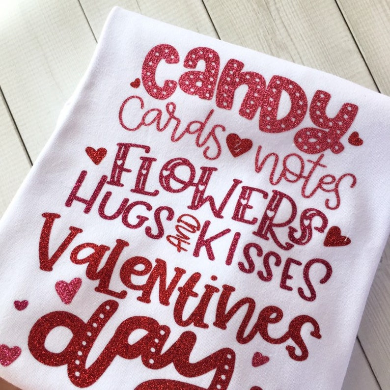 May include: White t-shirt with red glitter text that reads "candy, cards & notes, flowers, hugs & kisses, valentines day".