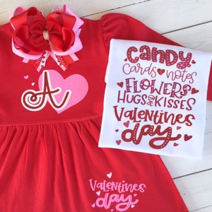 May include: Red and white Valentine's Day dress with a pink heart and the letter "A" on the front. The dress has a white long-sleeved shirt with the text "candy, cards notes, flowers, hugs & kisses, valentines day" on it.