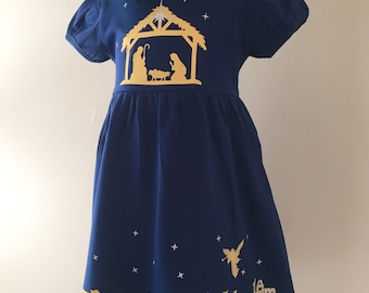 Könnte beinhalten: Marineblaues Kleid mit goldener Krippenszene, Sternen und dem Text "Come Let Us Adore Him". Das Kleid hat kurze Ärmel, einen Rüschensaum und eine große Schleife. Ein festliches Outfit für die Weihnachtszeit.