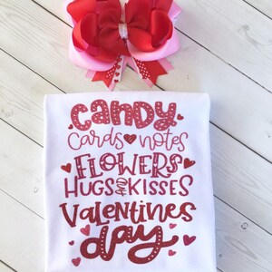 May include: White t-shirt with red glitter text that says "candy, cards, notes, flowers, hugs & kisses, valentines day". The shirt is decorated with small red hearts. A pink and red bow is in the background.
