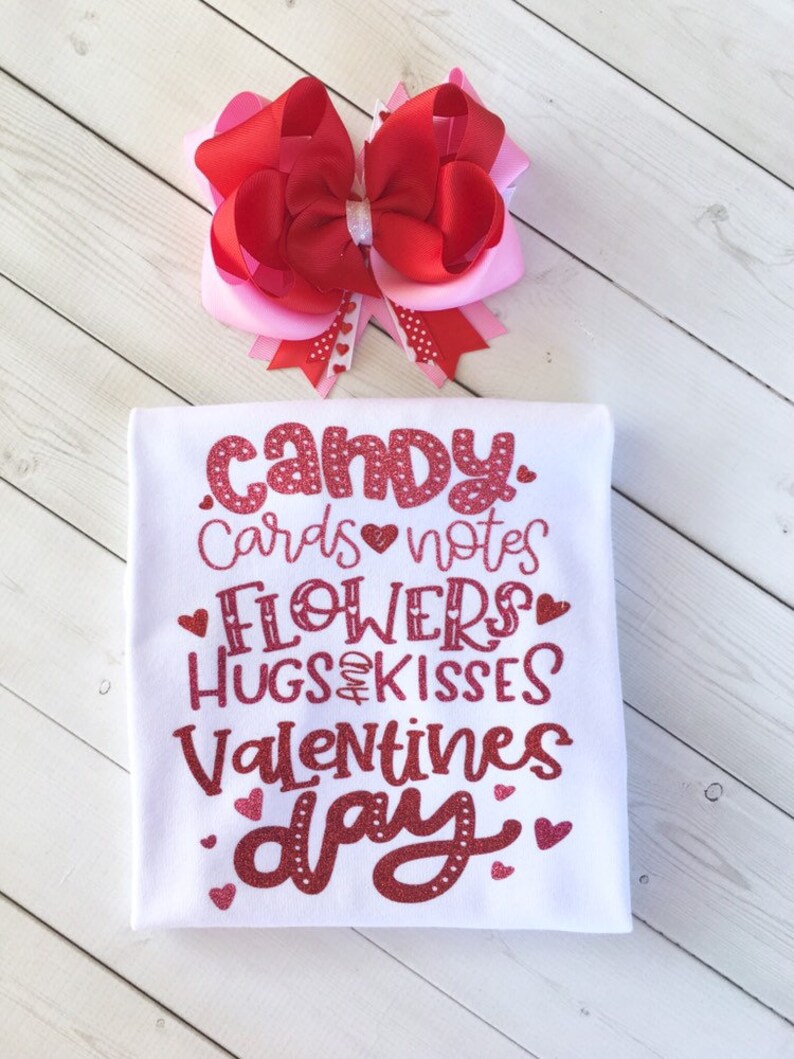 May include: White t-shirt with red glitter text that says "candy, cards, notes, flowers, hugs & kisses, valentines day". The shirt is decorated with small red hearts. A pink and red bow is in the background.