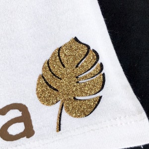 Może przedstawiać: Biała koszulka z nadrukiem złotej, błyszczącej liści monstera. Liść jest obwiedziony czarną linią. Na koszulce nadrukowane jest słowo "Aloha" w kolorze brązowym.