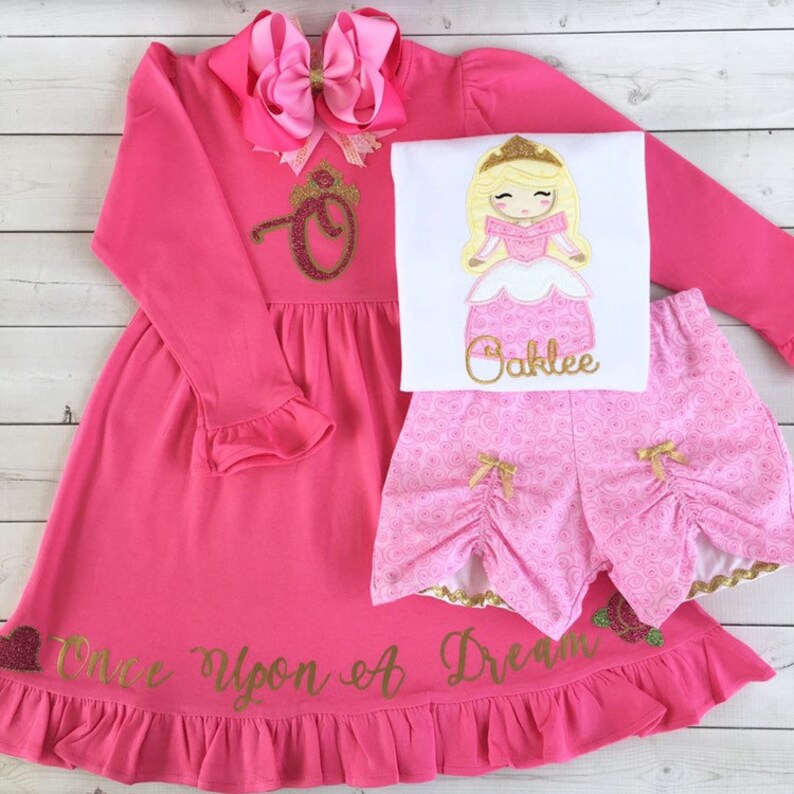 Peut inclure: Une robe rose &agrave; manches longues avec une lettre O en paillettes dor&eacute;es surmont&eacute;e d'une couronne. La robe est orn&eacute;e d'une inscription en paillettes dor&eacute;es qui dit "Once Upon A Dream". La robe est associ&eacute;e &agrave; un t-shirt blanc avec un personnage de princesse en paillettes dor&eacute;es et le nom "Oaklee" brod&eacute; dessus. La tenue est compl&eacute;t&eacute;e par un short rose avec une bordure en paillettes dor&eacute;es et un n&oelig;ud rose et dor&eacute;.