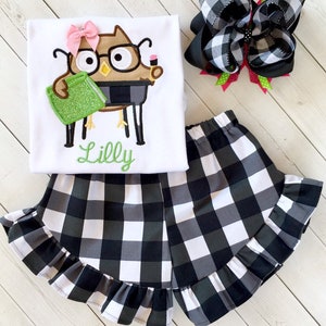 May include: A white t-shirt with an embroidered owl wearing glasses and holding a book. The shirt has the name "Lilly" embroidered below the owl. The shirt is paired with black and white checkered shorts with a ruffled hem. A black and white checkered bow is also included.