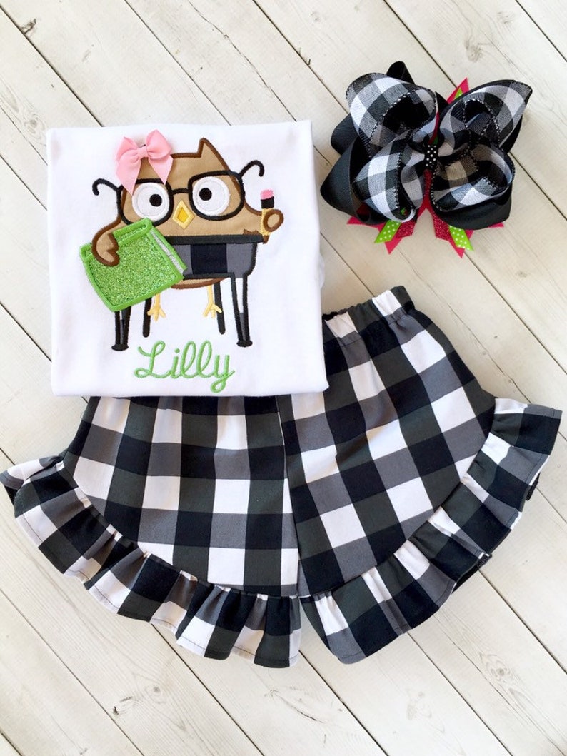 May include: A white t-shirt with an embroidered owl wearing glasses and holding a book. The shirt has the name "Lilly" embroidered below the owl. The shirt is paired with black and white checkered shorts with a ruffled hem. A black and white checkered bow is also included.