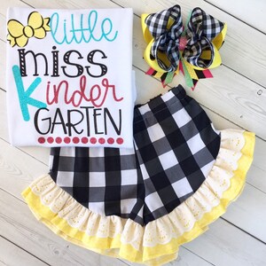 Pu&ograve; includere: Una maglietta bianca con il testo "little miss kindergarten" in lettere colorate. La maglietta &egrave; abbinata a pantaloncini a quadri neri e bianchi con un bordo di volant giallo e un fiocco per capelli a quadri giallo e nero.