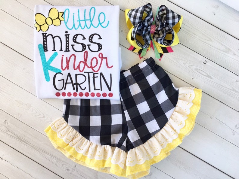 Pu&ograve; includere: Una maglietta bianca con il testo "little miss kindergarten" in lettere colorate. La maglietta &egrave; abbinata a pantaloncini a quadri neri e bianchi con un bordo di volant giallo e un fiocco per capelli a quadri giallo e nero.