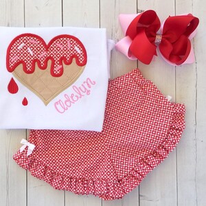 May include: A white onesie with a red heart-shaped ice cream cone design and the name "Adelyn" embroidered in pink. The onesie is paired with red and white heart-patterned bloomers and a pink and red bow.