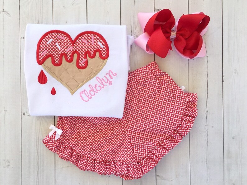 May include: A white onesie with a red heart-shaped ice cream cone design and the name "Adelyn" embroidered in pink. The onesie is paired with red and white heart-patterned bloomers and a pink and red bow.