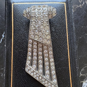 May include: A silver-toned metal brooch featuring a geometric, tie-like shape encrusted with numerous clear, round-cut rhinestones. The design includes a solid upper section and a lower portion composed of five vertical, rhinestone-lined bars.