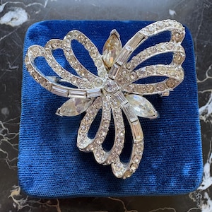 May include: A silver-toned metal brooch shaped like a stylised bow or butterfly, featuring multiple clear round rhinestones and elongated baguette-cut crystals. The piece is accented with a single amber-coloured marquise-cut stone at the top centre.