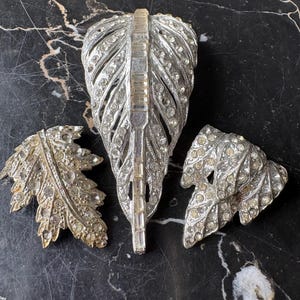 Op de afbeelding: Drie vintage broches op een zwart marmeren oppervlak. De grootste is een zilverkleurige veer, bezet met heldere strass-steentjes. Twee kleinere blad-vormige broches met vergelijkbare strass-details maken de set compleet.