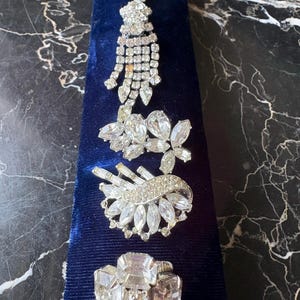 Peut inclure: Une collection de quatre broches en métal argenté ornées de strass transparents à facettes. Les pièces comprennent un motif à pampilles pendantes, une composition florale, un motif en éventail avec des pierres baguette et une broche rectangulaire.