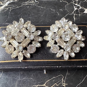 May include: A pair of circular clip-on earrings featuring a central square arrangement of clear baguette and round rhinestones, surrounded by a radiating border of marquise and round cut clear crystals set in a silver-toned metal frame.