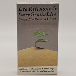May include: A compact disc case featuring the text "Lee Ritenour & Dave Grusin Live From The Record Plant." The cover art displays a small plant growing from sand. Additional text includes the names of the musicians.
