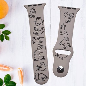 Può includere: Un cinturino per Apple Watch in silicone grigio con una stampa nera di gatti in varie pose di yoga. Il cinturino è mostrato su uno sfondo bianco.