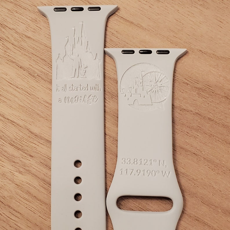 Pode incluir: Duas bandas brancas para Apple Watch com designs gravados. A banda da esquerda apresenta uma silhueta do castelo de Cinderela e o texto "It all started with a "magic" ". A banda da direita apresenta uma silhueta do castelo de Cinderela e as coordenadas "33.8121&deg; N, 117.9190&deg; W".