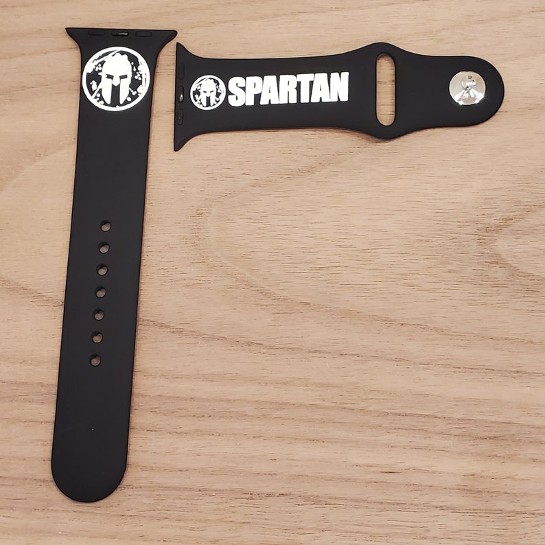 Pode incluir: Pulseira de rel&oacute;gio Apple de silicone preto com um logotipo Spartan branco e uma fivela prateada.
