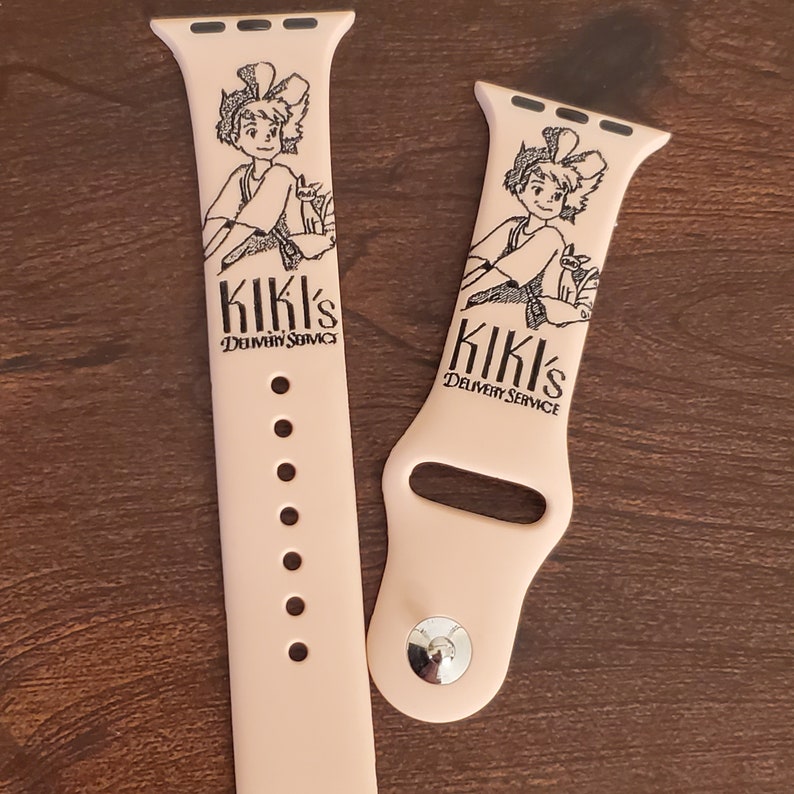 Pode incluir: Duas pulseiras de silicone rosa para Apple Watch com uma imagem em preto e branco de uma menina com um gato, e o texto "Kiki's Delivery Service".