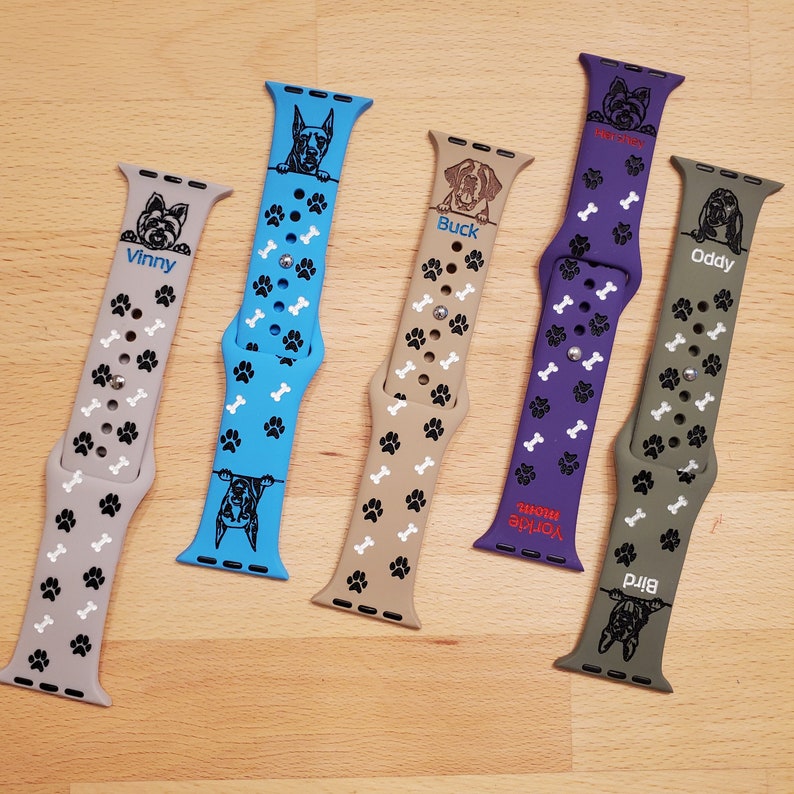 May include: Five different Apple Watch bands with dog breeds printed on them. The bands are in various colors, including blue, purple, green, and beige. Each band has a different dog breed and name printed on it, including Vinny, Buck, Oddy, Birda, and Yorkie Mom.